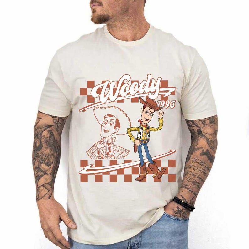 May include: Cream-colored t-shirt featuring a graphic of Woody from Toy Story. The design includes the word "Woody" above a checkered pattern and the year "1995". Woody is depicted in his cowboy attire, complete with a hat and boots.