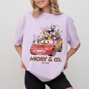 May include: A light purple t-shirt featuring a red race car with Disney characters, including Mickey Mouse, Donald Duck, and Goofy. The text "MICKEY & CO. EST 1928" is printed in gold.