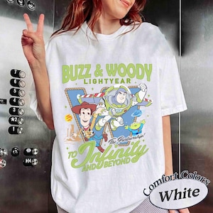 Peut inclure: Un t-shirt blanc &agrave; manches courtes avec un motif repr&eacute;sentant Buzz l'&Eacute;clair, Woody et un extraterrestre vert. Le texte indique BUZZ & WOODY LIGHTYEAR et TO INFINITY AND BEYOND. Le design inclut un fond triangulaire bleu et des &eacute;toiles jaunes.