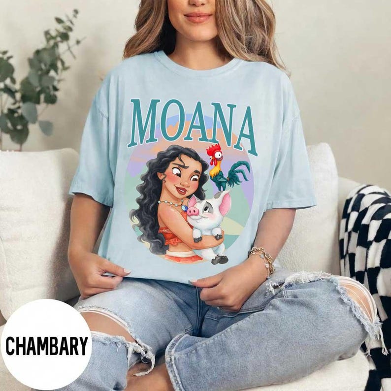 May include: Light blue t-shirt featuring the word "MOANA" in teal lettering. The shirt displays a colorful graphic of Moana holding a pig, with a rooster beside them. The shirt is made of a soft material.