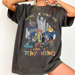 Könnte beinhalten: Dunkelgraues T-Shirt mit einer farbenfrohen Grafik von Zeichentrickfiguren und dem Text "WHERE ANYONE CAN BE ANYTHING". Das Design umfasst eine Stadtlandschaft und verschiedene Tierfiguren in Polizeiuniformen und Freizeitkleidung.