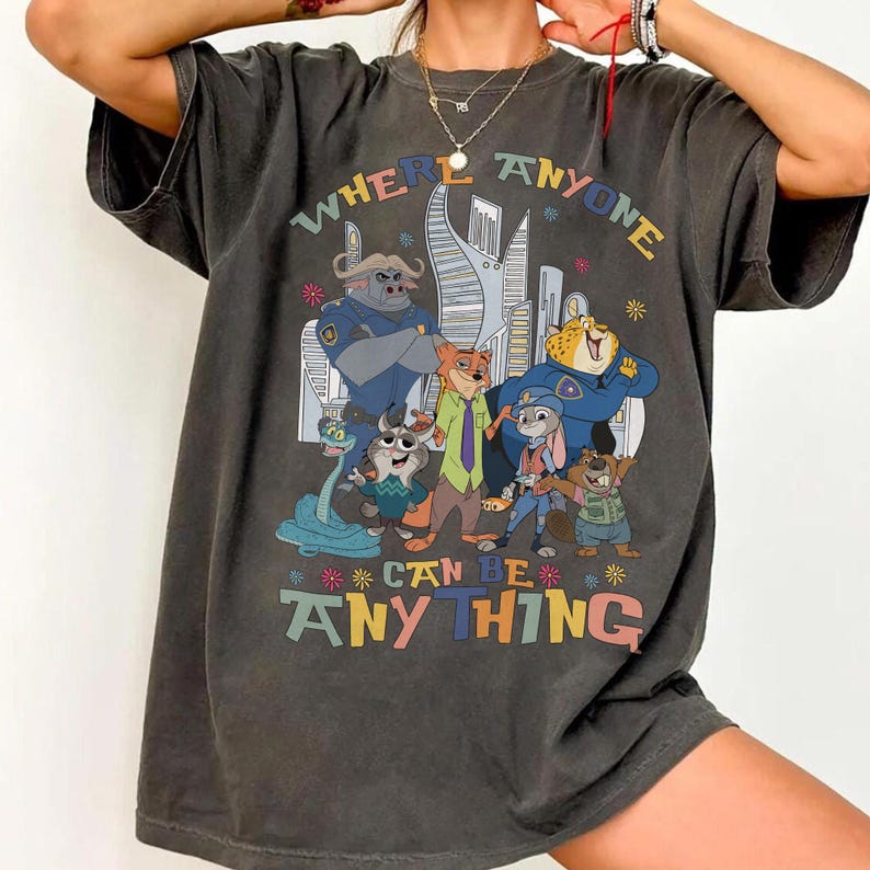 Puede incluir: Camiseta gris oscuro con un gr&aacute;fico colorido de personajes de dibujos animados y el texto "WHERE ANYONE CAN BE ANYTHING". El dise&ntilde;o incluye un paisaje urbano y varios personajes de animales con uniformes de polic&iacute;a y ropa informal.