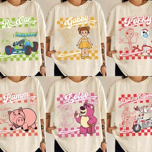 May include: Cream-colored t-shirts featuring various cartoon characters and text. Designs include a race car, a doll, a fork and spoon, a pig, a bear, and a motorcycle rider. Each design is set against a checkered background.