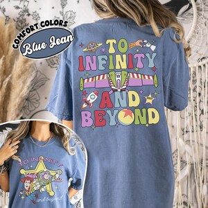 May include: Blue jean Comfort Colors t-shirt with a colorful graphic that says "To Infinity and Beyond." The design includes a rocket ship, a planet, and a space shuttle. The shirt also has a smaller graphic on the front.