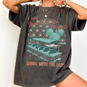 Könnte beinhalten: Dunkelgraues T-Shirt mit einer Bootszene und dem Text "Only Then Will We Truly Be Living With The Land". Das Design umfasst ein Boot auf dem Wasser, geometrische Formen und stilisierte Bäume. Das T-Shirt hat einen Vintage-Look.