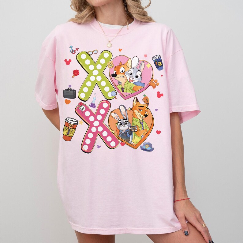 May include: Pink t-shirt featuring a colorful graphic of the characters Nick Wilde and Judy Hopps from Zootopia. The design includes the letters "X" and "O" with heart-shaped frames, coffee cups, and other themed elements.