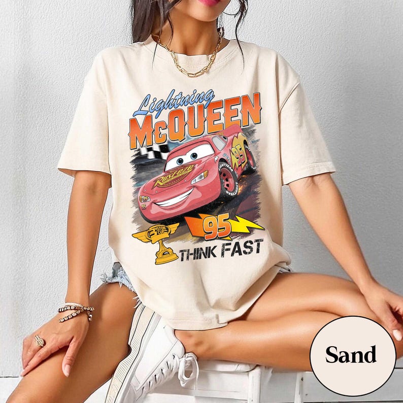 May include: Sand-colored t-shirt featuring a graphic of Lightning McQueen from the Cars movie. The design includes the text "Lightning McQueen," "95," and "Think Fast." The shirt has short sleeves and a crew neck.