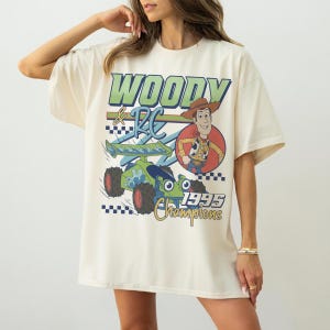May include: Cream-colored t-shirt featuring a graphic of Woody and RC from Toy Story. The design includes the text "WOODY & RC" in green and blue, a cartoon image of Woody, and a green and blue race car with the text "1995 Champions".