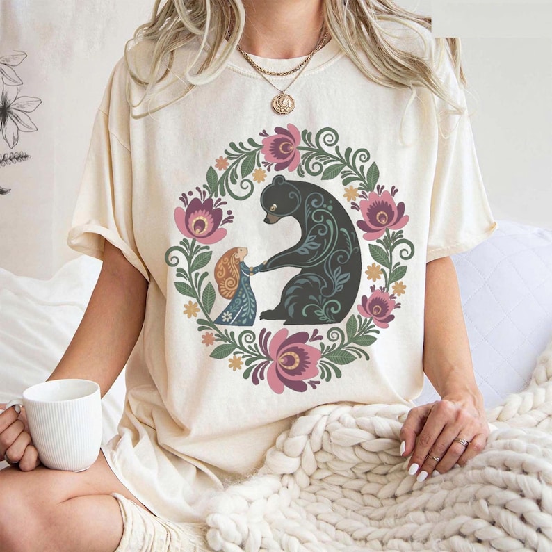 May include: Cream-colored t-shirt featuring a floral wreath design with a black bear and a girl holding hands. The wreath includes pink and purple flowers with green leaves. The bear has decorative patterns.