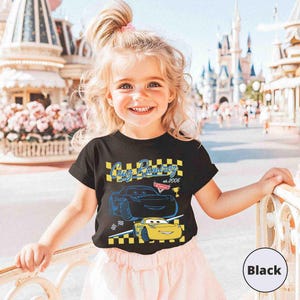 May include: A black t-shirt with a graphic of two Cars characters, Cruz Ramirez and a yellow car, with the text "Cruz Ramirez" and "Cars". The shirt is worn by a young child. The background includes a castle and other buildings.