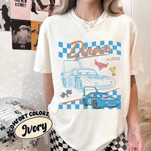 May include: Ivory t-shirt with a blue race car graphic and the text "Dinoco est. 2006" in orange and blue. The design includes a checkered flag and a trophy. The shirt also has the words "Comfort Colors Ivory" printed on it.