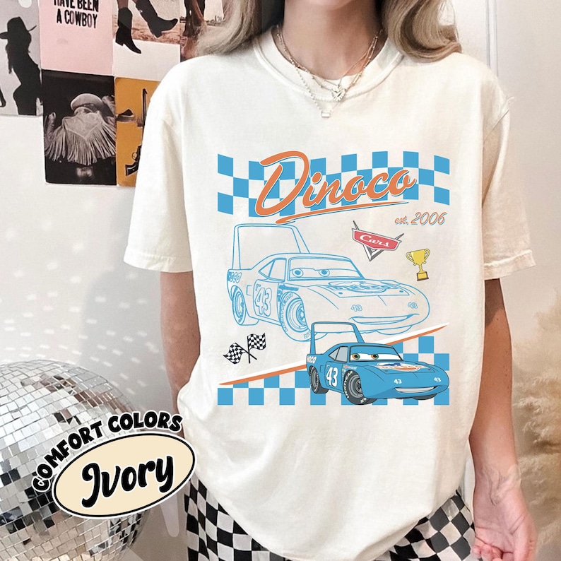 May include: Ivory t-shirt with a blue race car graphic and the text "Dinoco est. 2006" in orange and blue. The design includes a checkered flag and a trophy. The shirt also has the words "Comfort Colors Ivory" printed on it.