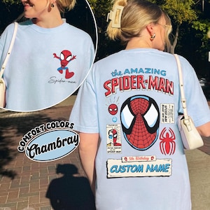 May include: Light blue t-shirt featuring Spider-Man graphics. The front has a small Spider-Man graphic and the text "Spider-man." The back displays "the AMAZING SPIDER-MAN" with a large Spider-Man mask design, additional graphics, and the text "CUSTOM NAME."