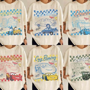 May include: Cream-coloured t-shirts featuring characters and designs from the animated film "Cars." Each shirt displays a character's name, a car illustration, and the year "2006" or "2005" in a retro style. The designs include blue, red, yellow, and green accents.