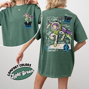 K&ouml;nnte beinhalten: Hellgr&uuml;nes T-Shirt mit Buzz Lightyear aus Toy Story. Die Vorderseite hat eine kleine Grafik, w&auml;hrend die R&uuml;ckseite ein gr&ouml;&szlig;eres Design mit dem Text "Toy Story", "Galactic Hero!" und "Space Ranger" zeigt. Das Shirt ist im Comfort-Colors-Stil.