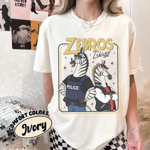 May include: Ivory t-shirt with a cartoon design of two zebras. One zebra is in a police vest, the other in a jacket. The shirt has the text "ZEBROS" in yellow and the words "Comfort Colors" and "Ivory" in a circular design.