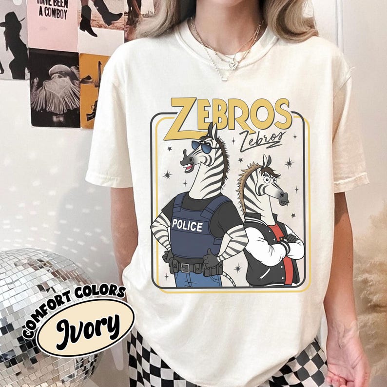 May include: Ivory t-shirt with a cartoon design of two zebras. One zebra is in a police vest, the other in a jacket. The shirt has the text "ZEBROS" in yellow and the words "Comfort Colors" and "Ivory" in a circular design.