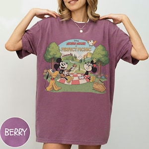 May include: Berry-colored t-shirt featuring a vintage-style graphic of Mickey Mouse, Minnie Mouse, and Pluto having a picnic. The graphic includes the text "Mickey Mouse PERFECT PICNIC". The shirt has short sleeves and a crew neck.