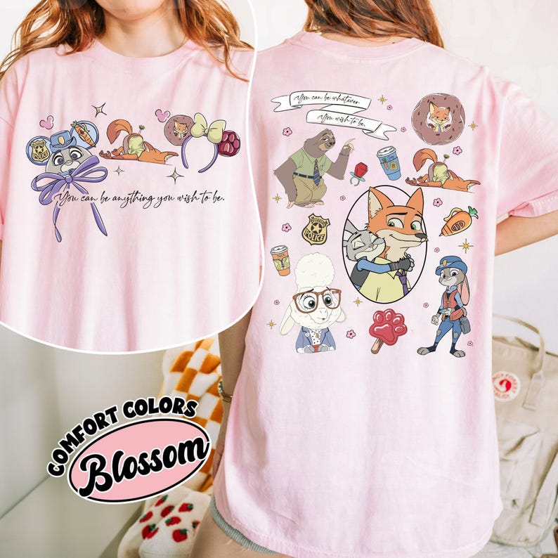 Puede incluir: Camiseta rosa claro con personajes de dibujos animados y la frase "You can be anything you wish to be." El dise&ntilde;o incluye un zorro, un conejo, un perezoso y otros personajes. La camiseta est&aacute; etiquetada como "Comfort Colors Blossom."