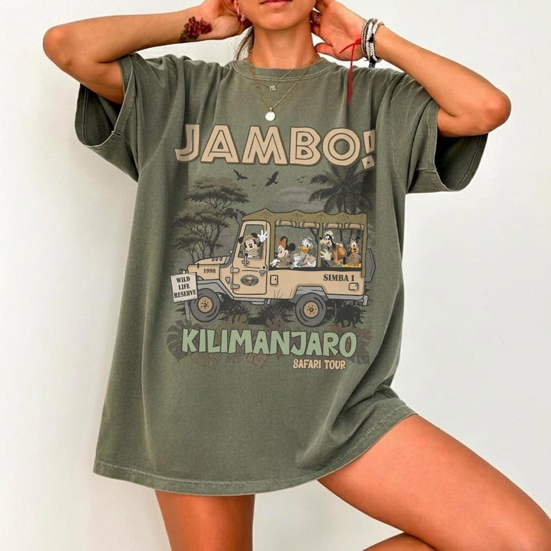May include: Olive green t-shirt featuring a safari scene with Mickey Mouse, Donald Duck, and other characters in a jeep. The shirt has the text "JAMBO!" and "KILIMANJARO SAFARI TOUR" in a vintage style.