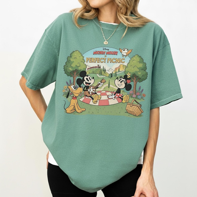 May include: A green t-shirt featuring a graphic of Mickey Mouse, Minnie Mouse, and Pluto having a picnic. The graphic includes the text "Mickey Mouse Perfect Picnic" and a scenic background with trees, flowers, and a bird.