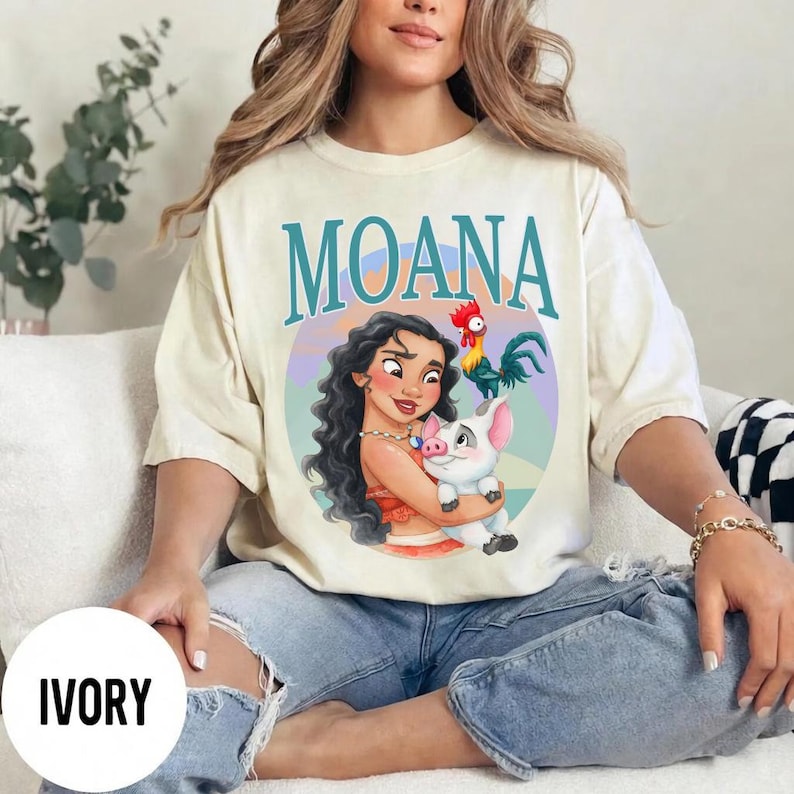 May include: Ivory-colored t-shirt featuring the word "MOANA" in teal letters. The shirt displays a colorful illustration of Moana holding a pig, with a rooster nearby. The shirt is made of a soft material.