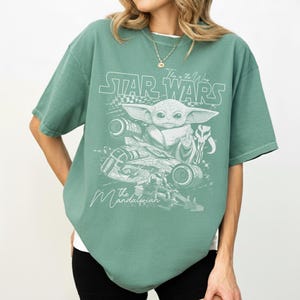 Puede incluir: Una camiseta de manga corta color verde salvia con un estampado gr&aacute;fico blanco de Grogu, una nave espacial y un casco mandaloriano. El texto dice Star Wars y The Mandalorian en una fuente estilizada. Tiene cuello redondo y un corte holgado.