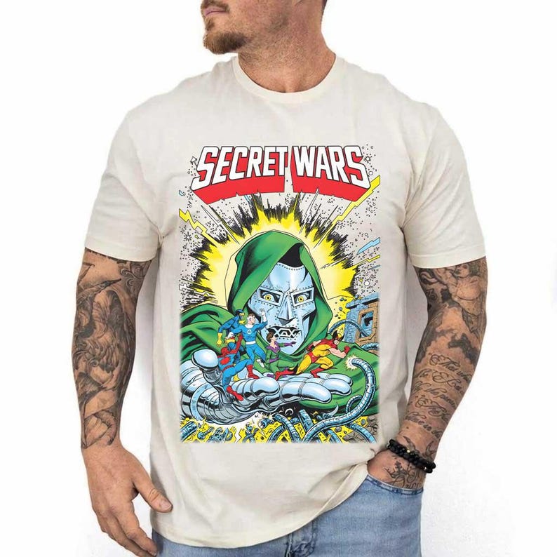 May include: Cream-colored t-shirt featuring a comic book graphic. The graphic includes the text "SECRET WARS" in red and white, and a depiction of a character with a green hood and silver mask, holding other comic book characters.
