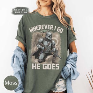 Puede incluir: Una camiseta de manga corta color verde musgo con un estampado gr&aacute;fico de un personaje con armadura que sostiene un rifle con una peque&ntilde;a criatura sobre el hombro. El texto dice WHEREVER I GO HE GOES. La camiseta tiene cuello redondo y corte holgado.