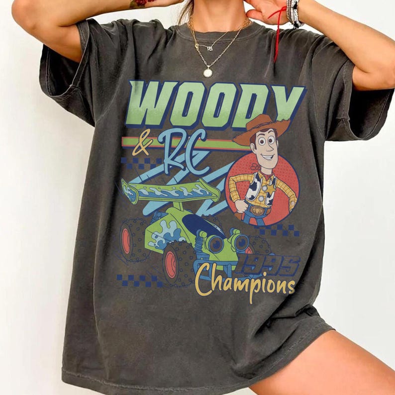 May include: Dark gray t-shirt featuring the text "WOODY & RC" above an illustration of Woody and a race car. The shirt also includes the text "1995 Champions" in a vintage style.