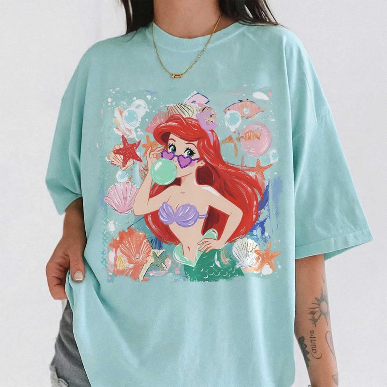 May include: A light blue t-shirt featuring a graphic print of a red-haired mermaid wearing heart-shaped sunglasses and blowing a green bubble. The design includes various seashells, starfish, and bubbles in a colorful, artistic style.