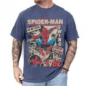 Pode incluir: Camiseta azul com um gr&aacute;fico vintage do Homem-Aranha. O design inclui as palavras "The Amazing Spider-Man", "New York" e "Swing Into Action", juntamente com o ano de "1962". O gr&aacute;fico mostra o Homem-Aranha em seu traje cl&aacute;ssico vermelho e azul.