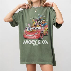 May include: Olive green t-shirt featuring a graphic of Disney characters riding in a red Lightning McQueen race car. The text "Mickey & Co. EST 1928" is printed below the car. The shirt has short sleeves and a relaxed fit.