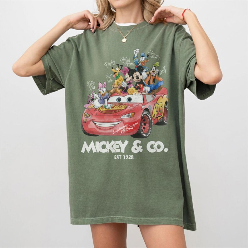 May include: Olive green t-shirt featuring a graphic of Disney characters riding in a red Lightning McQueen race car. The text "Mickey & Co. EST 1928" is printed below the car. The shirt has short sleeves and a relaxed fit.