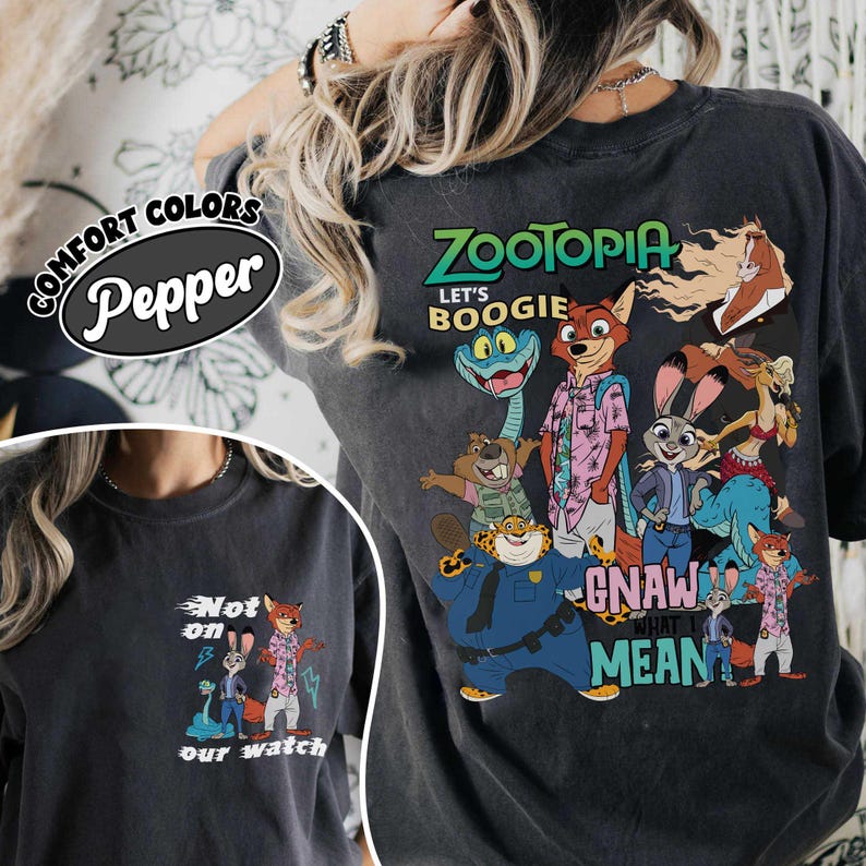 May include: Dark gray t-shirt featuring a colorful graphic of characters from the animated movie Zootopia. The design includes the text "Zootopia," "Let's Boogie," and "Gnaw What I Mean." The shirt also has a smaller graphic on the front.