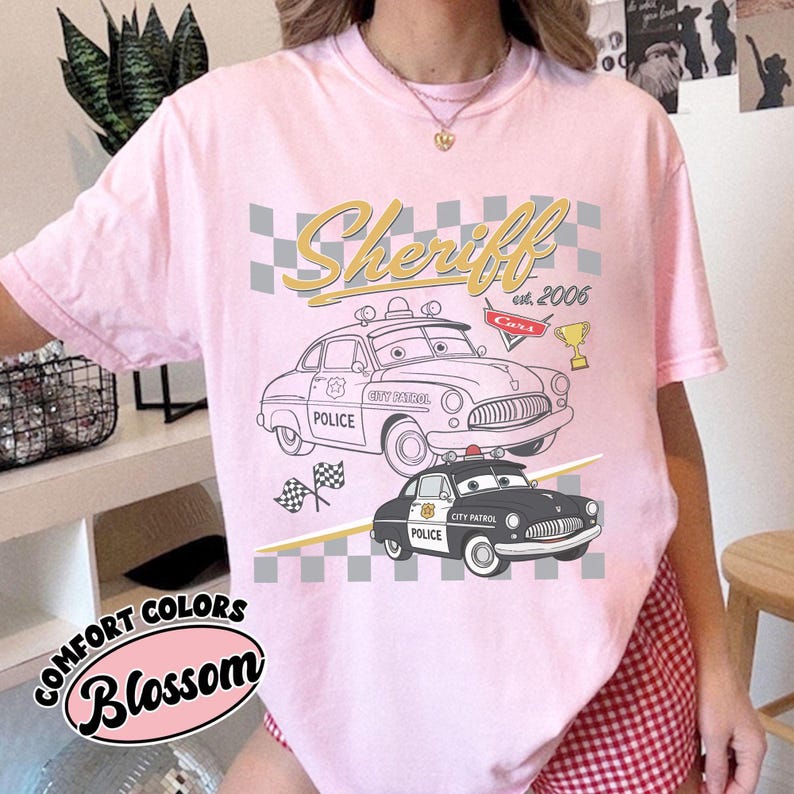 May include: A light pink Comfort Colors t-shirt with a graphic of two cartoon police cars and the text "Sheriff est. 2006 Cars". The shirt also has the text "Blossom" in a floral design.