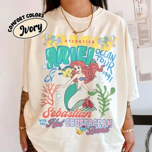 May include: An ivory t-shirt featuring a graphic print of Ariel, Flounder, and Sebastian. The text reads Atlantica Ocean Tour 1991, Ariel, Sebastian and the Hot Crustacean Band. The design includes coral, seaweed, and bubbles in blue, red, and yellow colors.