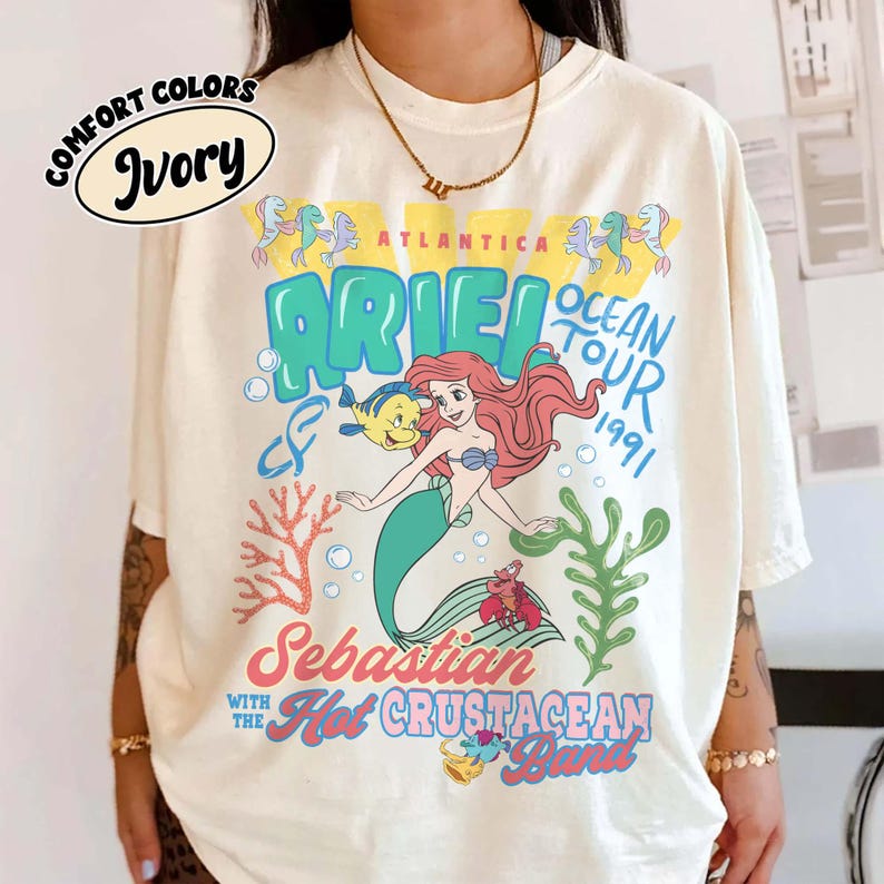 May include: An ivory t-shirt featuring a graphic print of Ariel, Flounder, and Sebastian. The text reads Atlantica Ocean Tour 1991, Ariel, Sebastian and the Hot Crustacean Band. The design includes coral, seaweed, and bubbles in blue, red, and yellow colors.