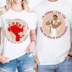 Puede incluir: Dos camisetas blancas con un diseño gráfico de un personaje de dibujos animados. La camiseta de la izquierda presenta un personaje de diablo rojo con un tridente y el texto "I'm gonna lead you down the path that rocks". La camiseta de la derecha presenta un personaje de ángel blanco con un arpa y el texto "I'm gonna lead you down the path of righteousness".