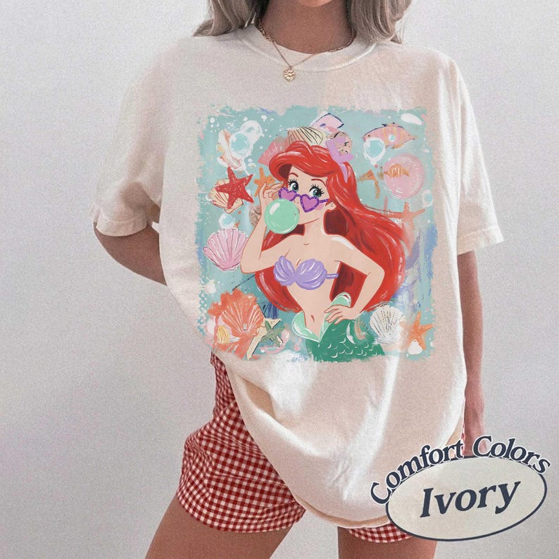 May include: An ivory t-shirt featuring a graphic print of a red-haired mermaid wearing heart-shaped sunglasses and blowing a bubble. The illustration includes various seashells and starfish in a light blue square frame on the front of the shirt.