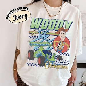 May include: Ivory-colored t-shirt featuring a graphic of Woody and RC from Toy Story. The design includes the text "WOODY & RC", a cartoon image of Woody, and a green and blue RC car. The shirt also has the text "1995 Champions".