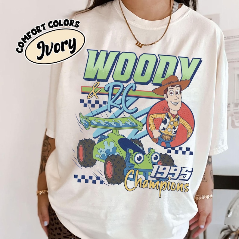 May include: Ivory-colored t-shirt featuring a graphic of Woody and RC from Toy Story. The design includes the text "WOODY & RC", a cartoon image of Woody, and a green and blue RC car. The shirt also has the text "1995 Champions".