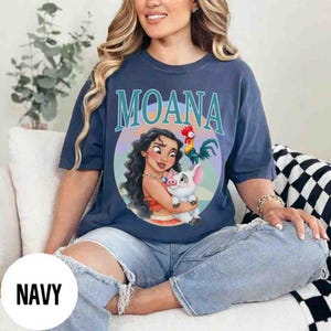 May include: Navy blue t-shirt featuring the word "MOANA" in teal lettering above a circular graphic. The graphic depicts a cartoon woman holding a pig, with a rooster to the right. The shirt is a relaxed fit.