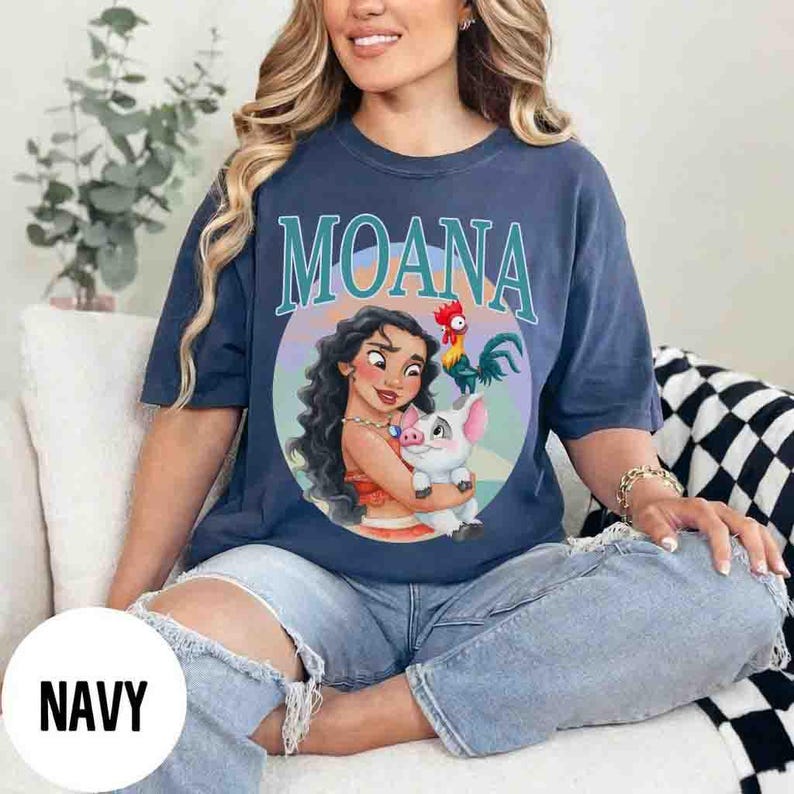 May include: Navy blue t-shirt featuring the word "MOANA" in teal lettering above a circular graphic. The graphic depicts a cartoon woman holding a pig, with a rooster to the right. The shirt is a relaxed fit.