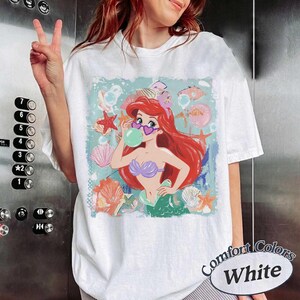 May include: A white short-sleeve t-shirt featuring a graphic print of a red-haired mermaid wearing heart-shaped sunglasses and blowing a green bubble. The design includes various seashells, starfish, and bubbles in shades of pink, orange, and blue.