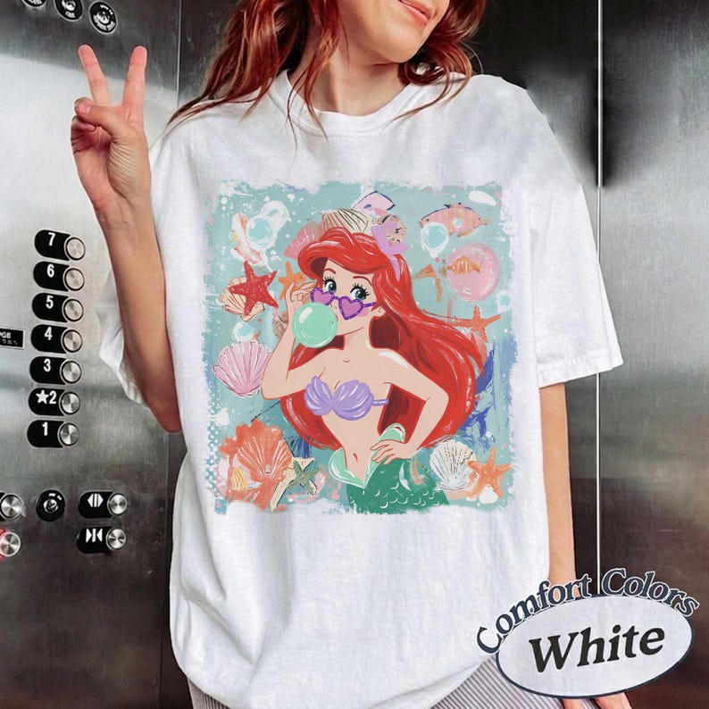 May include: A white short-sleeve t-shirt featuring a graphic print of a red-haired mermaid wearing heart-shaped sunglasses and blowing a green bubble. The design includes various seashells, starfish, and bubbles in shades of pink, orange, and blue.