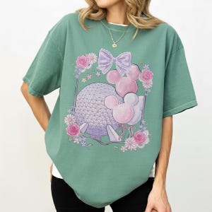 K&ouml;nnte beinhalten: Salbeigr&uuml;nes T-Shirt mit einer Grafik einer geometrischen Kugel, Mickey Mouse-Ballons und einer lila Schleife. Das Design ist von rosa Rosen und anderen Blumen umgeben. Das T-Shirt hat kurze &Auml;rmel und einen Rundhalsausschnitt.