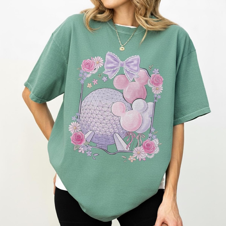 K&ouml;nnte beinhalten: Salbeigr&uuml;nes T-Shirt mit einer Grafik einer geometrischen Kugel, Mickey Mouse-Ballons und einer lila Schleife. Das Design ist von rosa Rosen und anderen Blumen umgeben. Das T-Shirt hat kurze &Auml;rmel und einen Rundhalsausschnitt.