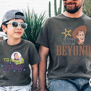 Puede incluir: Dos camisetas de color gris oscuro con gráficos de dibujos animados. Una camiseta presenta a Buzz Lightyear con "To Infinity" en escritura púrpura. La otra camiseta tiene a Woody con "And Beyond" en texto marrón. Ambas camisetas son usadas por personas.