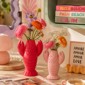 Könnte beinhalten: Zwei hummerförmige Vasen, eine leuchtend rosa und eine hellrosa, halten bunte Blumen. Die Vasen stehen auf einem Tisch mit Büchern, einer Kaffeetasse und einem Schild mit der Aufschrift "AMORE AMOUR AMOR LOVE". Im Hintergrund ein gerahmter "MATISSE"-Druck.